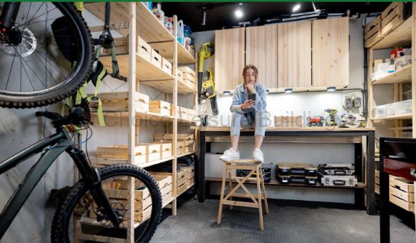 Home 40 A woman sitting on a stool in a garage workshop. The room is equipped with shelving and workbenches.