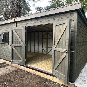 A wooden summerhouse with double doors and large windows, surrounded by trees and plants.