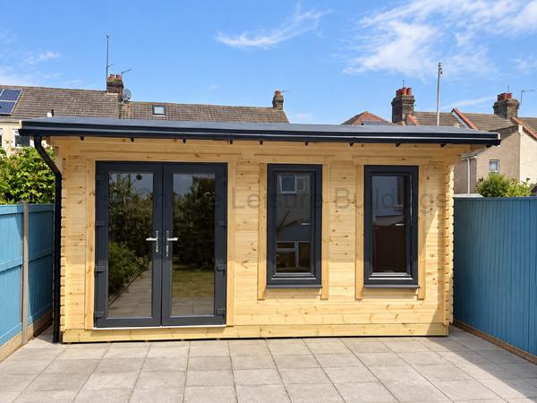 Pale timber log cabin with charcoal double doors and two tall windows, set on a tidy paved garden patio.