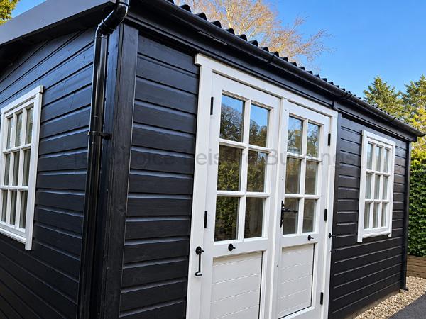 Small dark wooden garden room with white-framed doors and windows on a gravel lot.