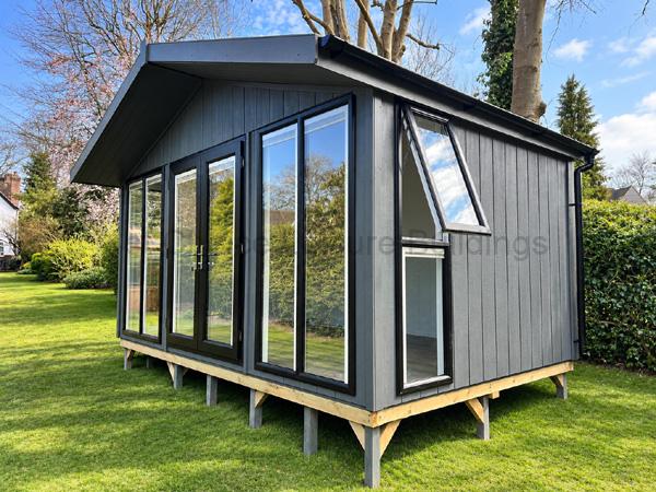Large dark grey wooden garden room on stilts with large glass doors and sloped roof in a grassy yard.