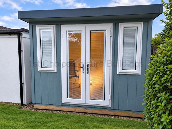 Small blue-grey Royal Buckingham external studio with white-framed doors, side windows, and lit interior.